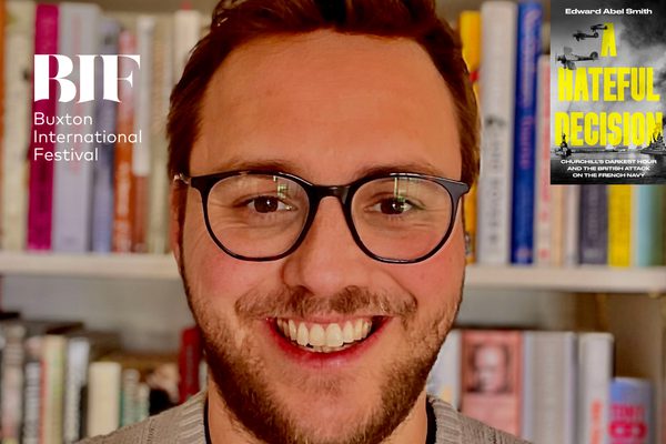 Man with brown hair and glasses smiles to camera, book shelves are visible behind him.