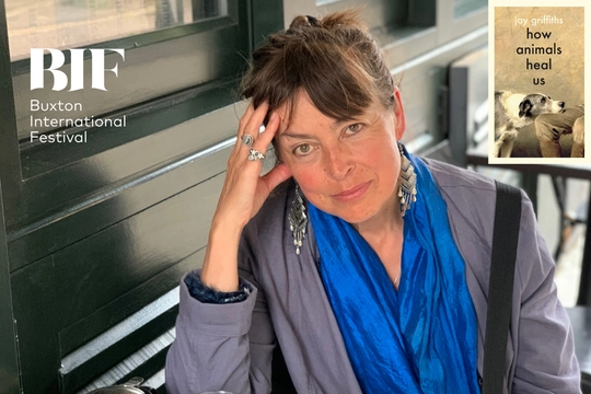 White woman with brunette fringe smiles at the camera. Her head resting on her silver ringed hand. She wears a blue top.