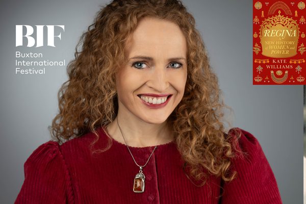 White woman with red curly hair smiles to the camera. She wears red lipstick and a red dress.