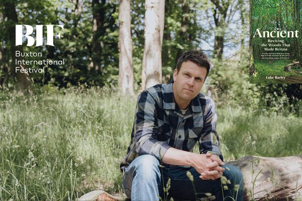White man with brunette hair sits on larger fallen tree. There is long grass behind him.