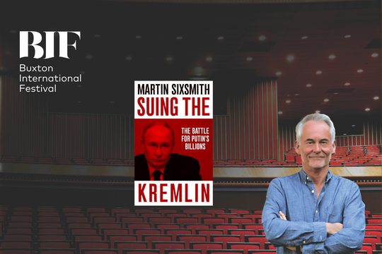 White man with white hair and stubble smile in a blue shirt. There are red theatre seats behind him.