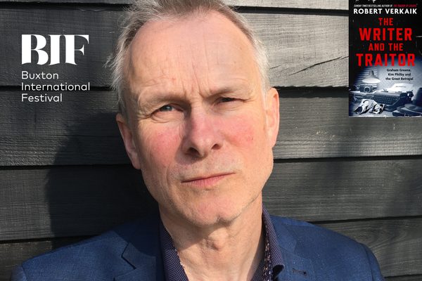 White man with white hair looks to camera, standing in front of dark wooden fence.