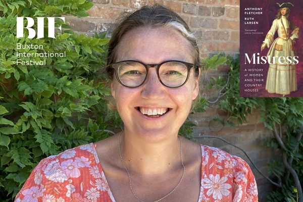 White woman with freckles and glasses smiles to camera. There is various green foliage behind her.