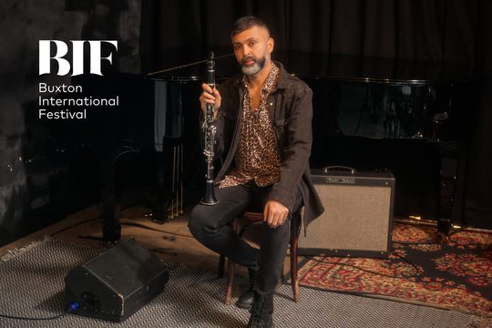 A man in a leopard print shirt and black jacket sits in front of a grand piano, holding a clarinet.