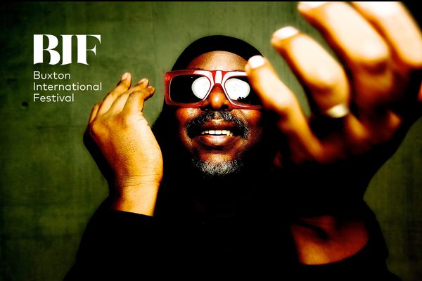 A man with a beard wearing wide-framed red sunglasses dances against a mossy green background.