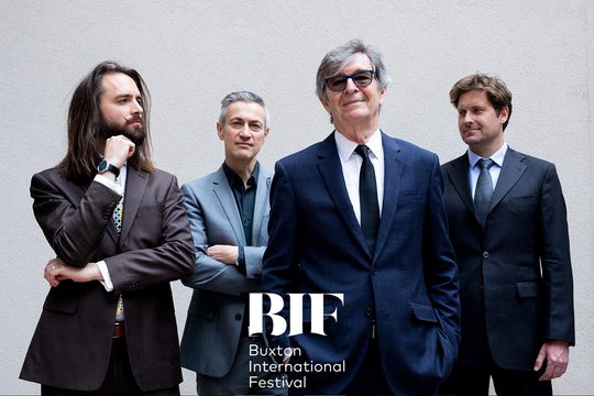 Four men in well-tailored suits in shades of blue pose in front of a grey background.