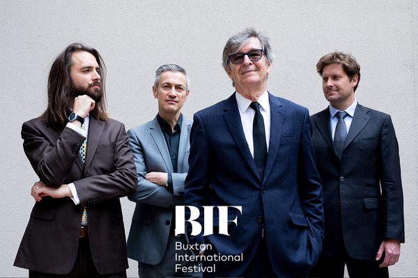 Four men in well-tailored suits in shades of blue pose in front of a grey background.