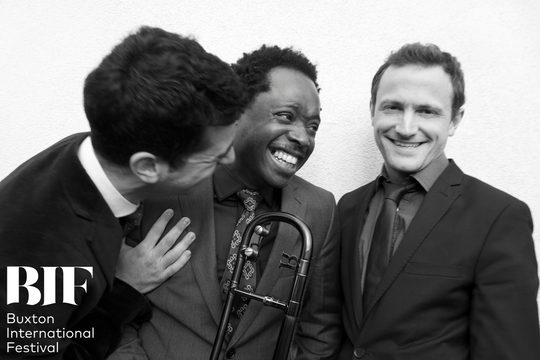 Black and white image of three men in suits laughing with each other. The man in the middle holds a trombone.