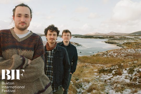 Three men in warm jumpers stand in front of a snowy lake.