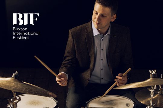 A man in a light shirt and dark brown jacket sits and plays the drums.