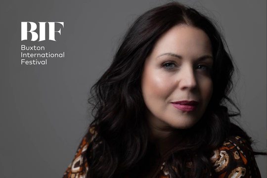 A woman with dark hair wearing a patterned top and pink lipstick sits in front of a grey background.