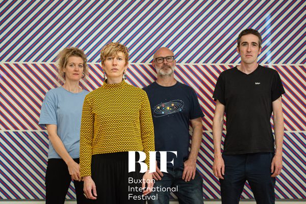Two blonde women in bright coloured tops, one blue and one yellow, and two men in dark t-shirts stand in front of a vibrant background of alternating diagonal stripes.
