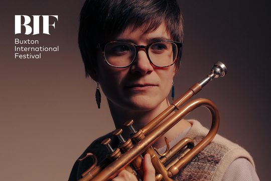 A woman with short hair and glasses wears a sweater vest and holds a trumpet.