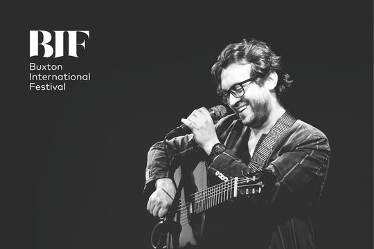 A black and white image of a man holding a guitar. He is standing at a microphone and smiling.