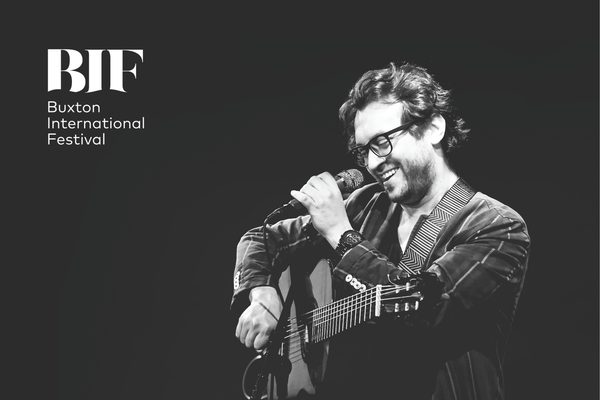 A black and white image of a man holding a guitar. He is standing at a microphone and smiling.