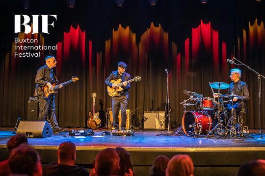 Three men in black suits perform on a stage. Two are playing guitars and one is playing drums.
