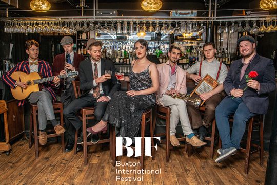 A group of 7 musicians all dressed up in suits and dresses sit on bar stools, sipping drinks and playing instruments.