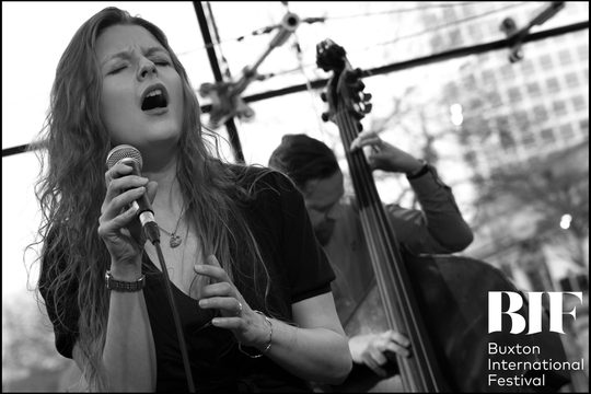 A black and white image of a woman with long hair singing into a microphone. She is standing in front of a man playing the double bass.