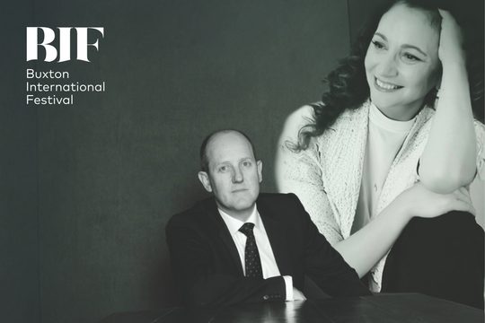 Woman with brunette curly hair smiles resting her elbow on her knee. She wears a pale shirt and jacket. Beside and image of a man smiling into the camera wearing a suit and tie.