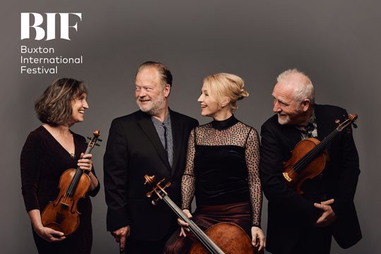 Four people dressed in black smile and laugh with each other holding various string instruments.