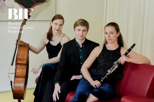 Three performers, two women and man sit of a red sofa wearing dark clothes. They smile to the camera