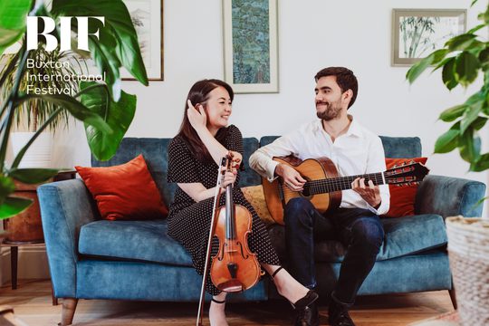 A woman and a man sit on a blue sofa. He plays the guitar while she is holding a violin.
