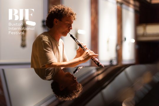 Man with curly hair and cream t-shirt plays the oboe.