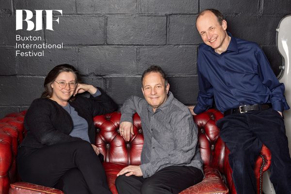 Three performers, one woman and two men, sit on a red leather sofa in front of a black brick wall.