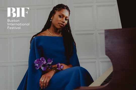 Black woman with long braids sits at a piano smiling. She wears a blue off the shoulder dress and holds a purple flower in her hand.