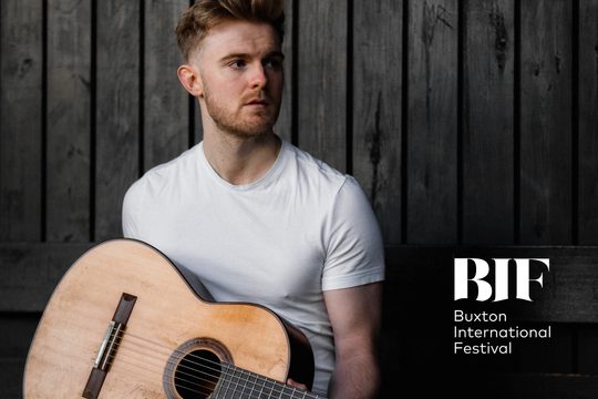 Young man with ginger hair and stubble holds a guitar. He is wearing a white t-shirt.