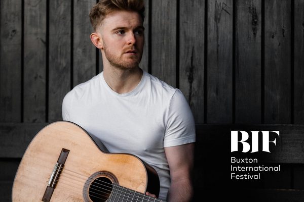 Young man with ginger hair and stubble holds a guitar. He is wearing a white t-shirt.