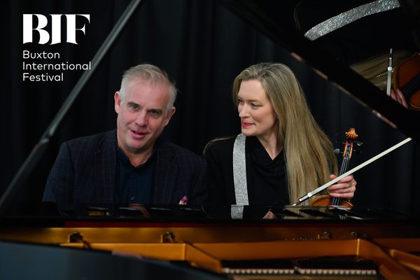 Man with grey hair sits at a piano, playing. A woman with long blonde hair sits beside him holding a violin on her knee.