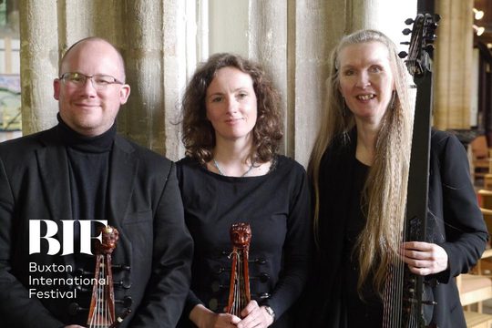 Three people a bald man with glasses, a brunette woman with curly hair and a blonde woman with long straight hair smile to the camera. The wear all black.