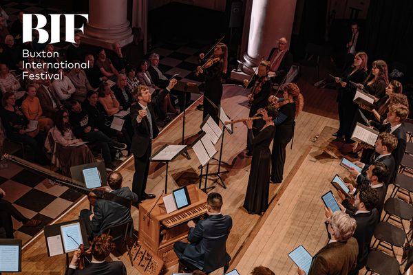 Man stands in front of orchestra conducting, while audience sit behind watching.