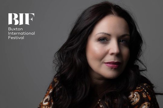 White woman with dark hair and bright pink lipstick looks to camera in front of grey background.