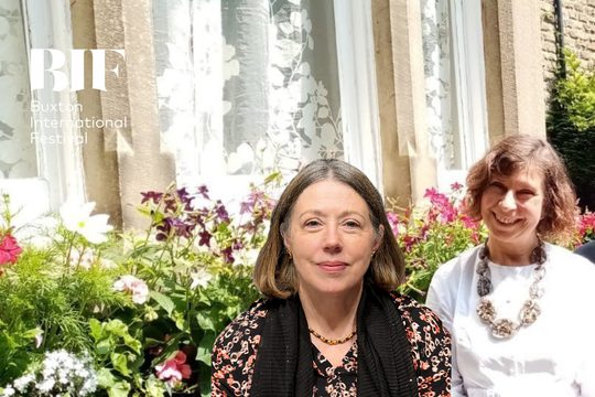 Two women with short brunette hair, one wears a white shirt, the other wears a floral blouse against a backdrop of greenery and flowers.