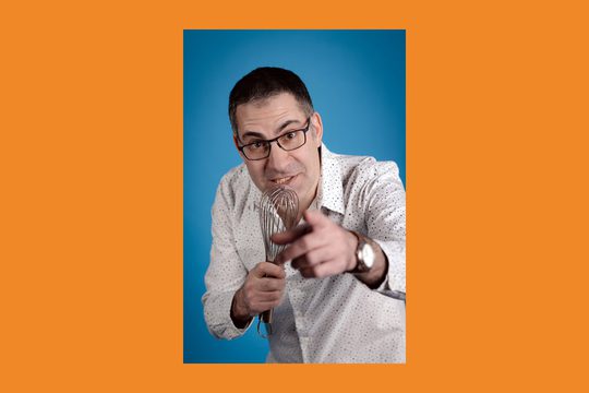 An orange background with an image of a man in a white spotty shirt talking into a whisk as if it was a microphone.