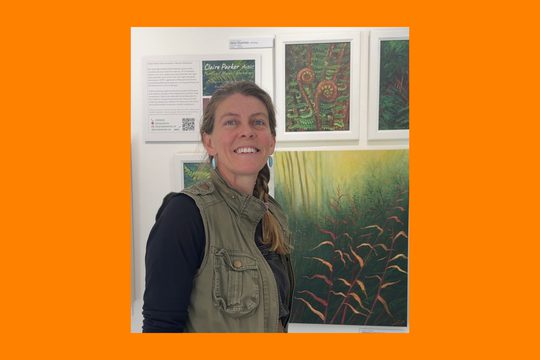 A woman wearing a green jacket. She has a plait in her hair and in standing in front of paintings of nature.