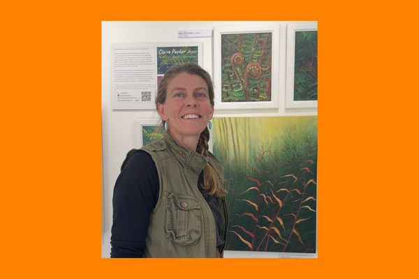 A woman wearing a green jacket. She has a plait in her hair and in standing in front of paintings of nature.