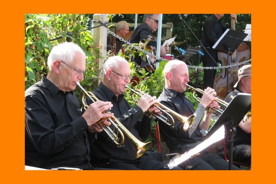Three white men wearing black suits play the trumpet in a garden.