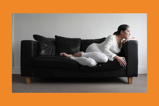 A white, brunette woman wearing all white lies on a black leather sofa.