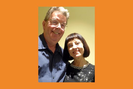 An older white man with grey hair wearing a blue shirt with an older woman with chin-length brown hair wearing a black dress.