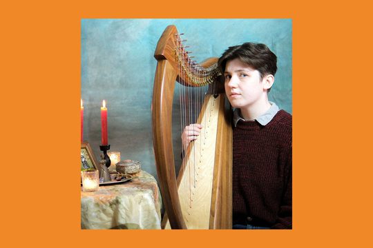 A performer with short brown hair wearing a red knitted jumper holds a harp.