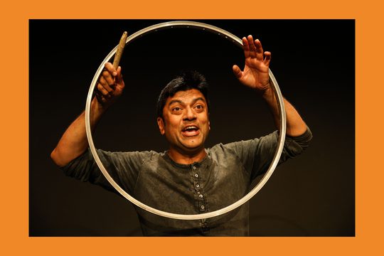 A man with dark hair wearing a long-sleeved grey t-shirt holds up a large metal circle around his face.