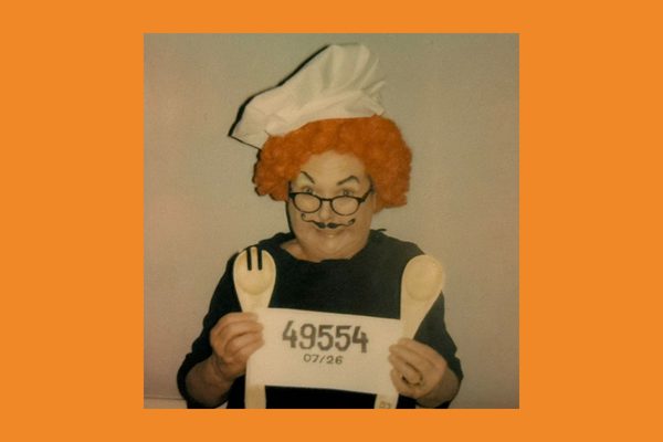 A man wearing an orange curly wig and a chef hat. He is holding up paper with numbers written on, and is holding up wooden cooking utensils.
