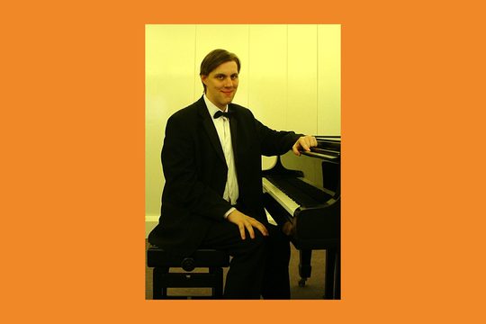 A man in a white shirt, black suit and bow tie sits at a grand piano and smiles.