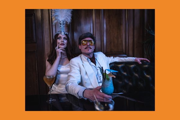 A man in a white suit wearing sunglasses sat on a sofa with a woman in a gold dress and feathered headdress. They are drinking cocktails.