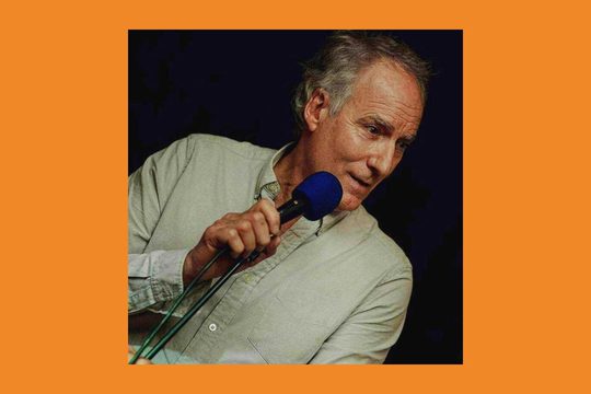 An older man with grey hair wearing a grey shirt holds a blue microphone to his mouth.