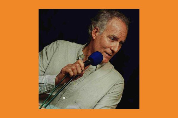 An older man with grey hair wearing a grey shirt holds a blue microphone to his mouth.