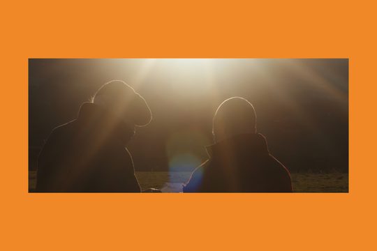 Two men in winter coats stand in a field with the sun beaming down on them.
